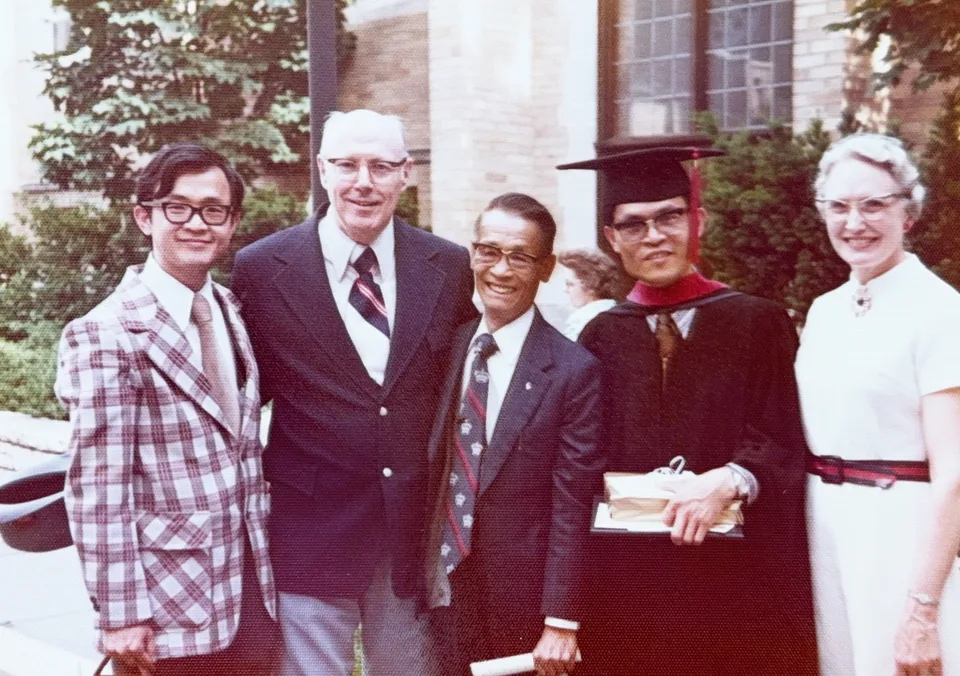 Colour photograph outside a stone building: five people stand together, David in academic gown and mortarboard holding his diploma, with his brother, his father and an older American couple.