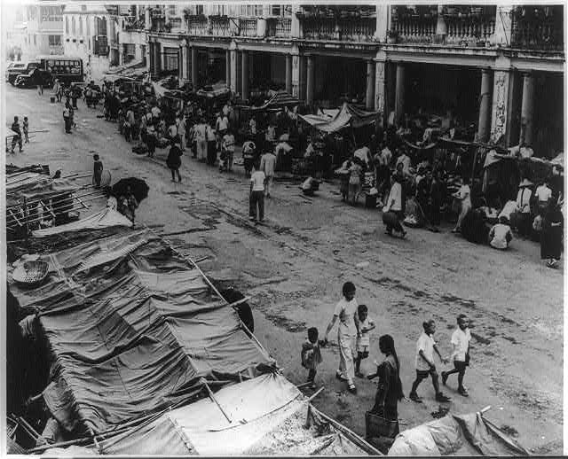 A crowded Kowloon street lined with food stalls and makeshift refugee huts, 1955.