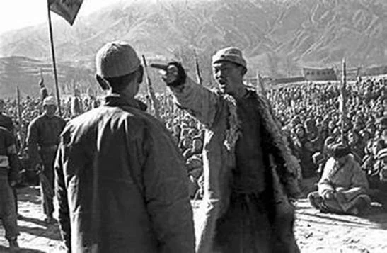 Black-and-white photograph: at an open-air meeting a man in a cap shouts and points at a man standing with his back to the camera, before a seated crowd under flags.
