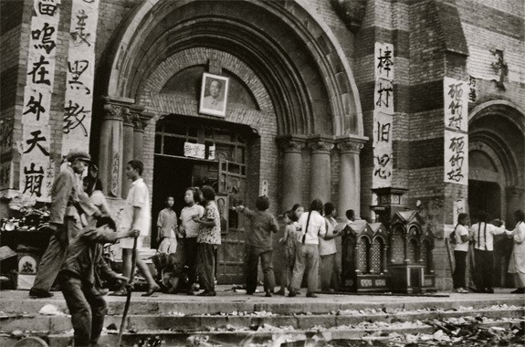 Black-and-white photograph: people outside a church doorway hung with a portrait of Mao and slogan banners, broken objects on the steps.