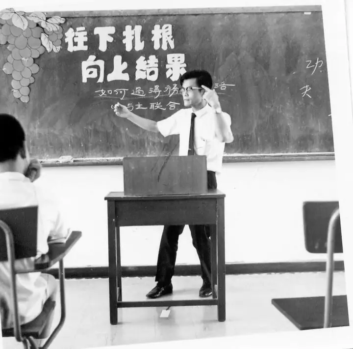 An archival photograph of a man teaching at a lectern before a Chinese chalkboard.