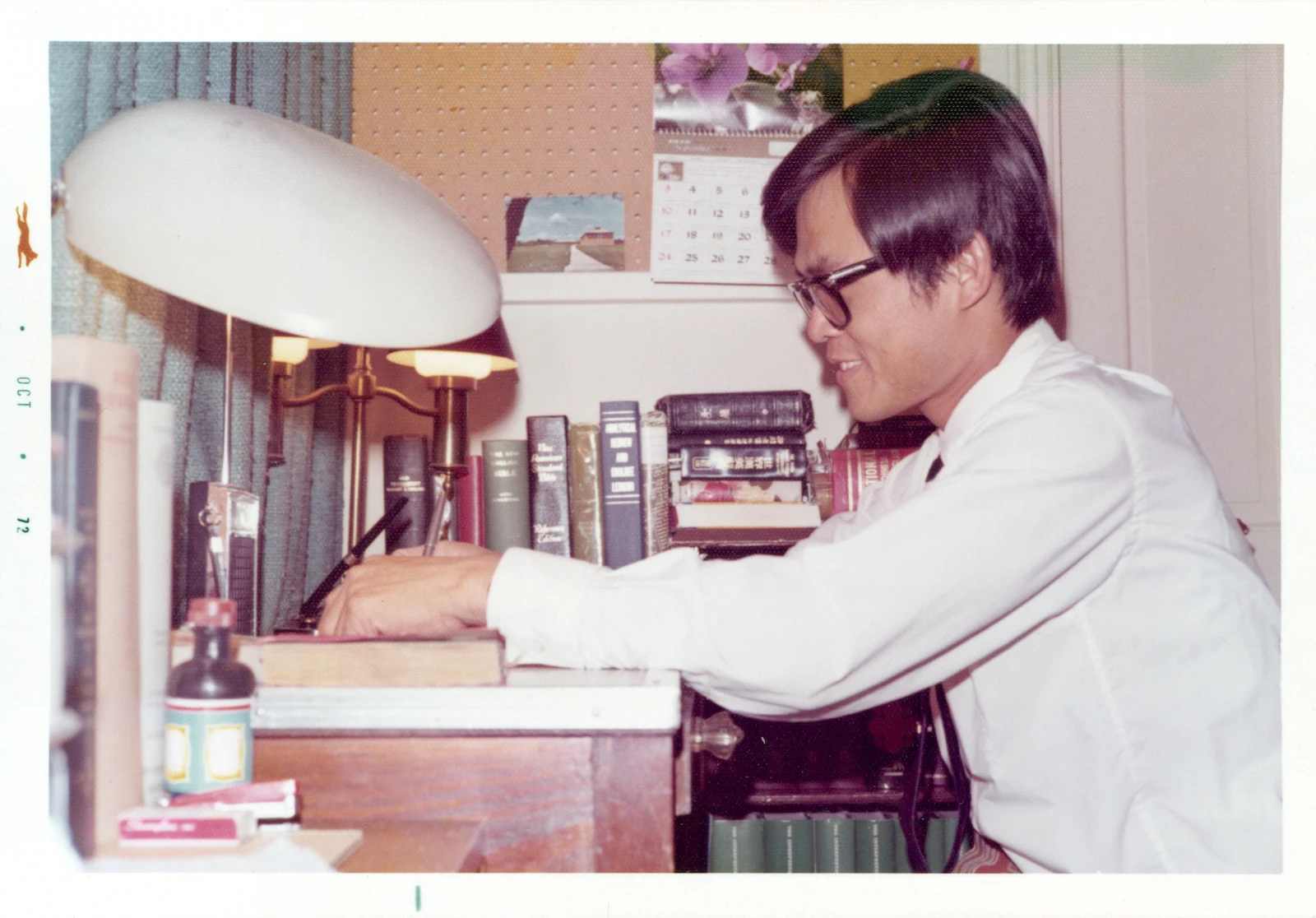 David, wearing glasses and a white shirt, writes at a desk crowded with books beneath a large lamp.