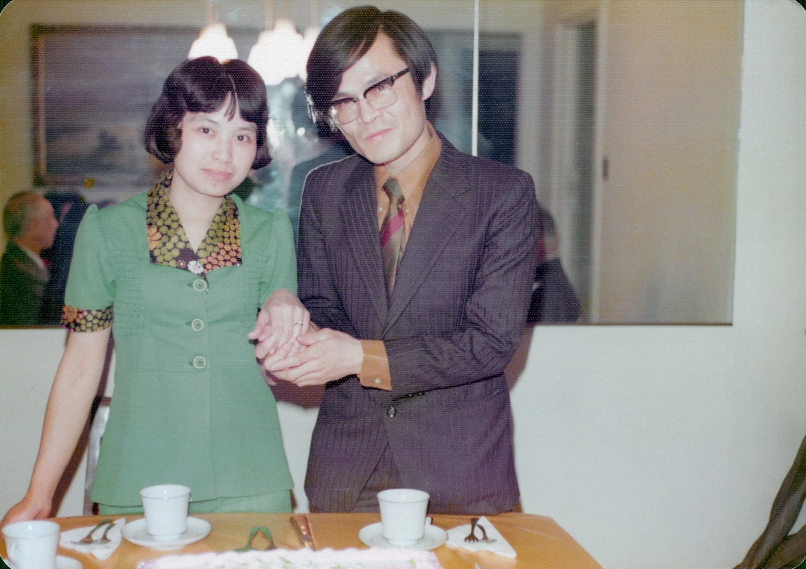 David supports Esther’s raised left hand to show her engagement ring as they stand behind a table set with cups.