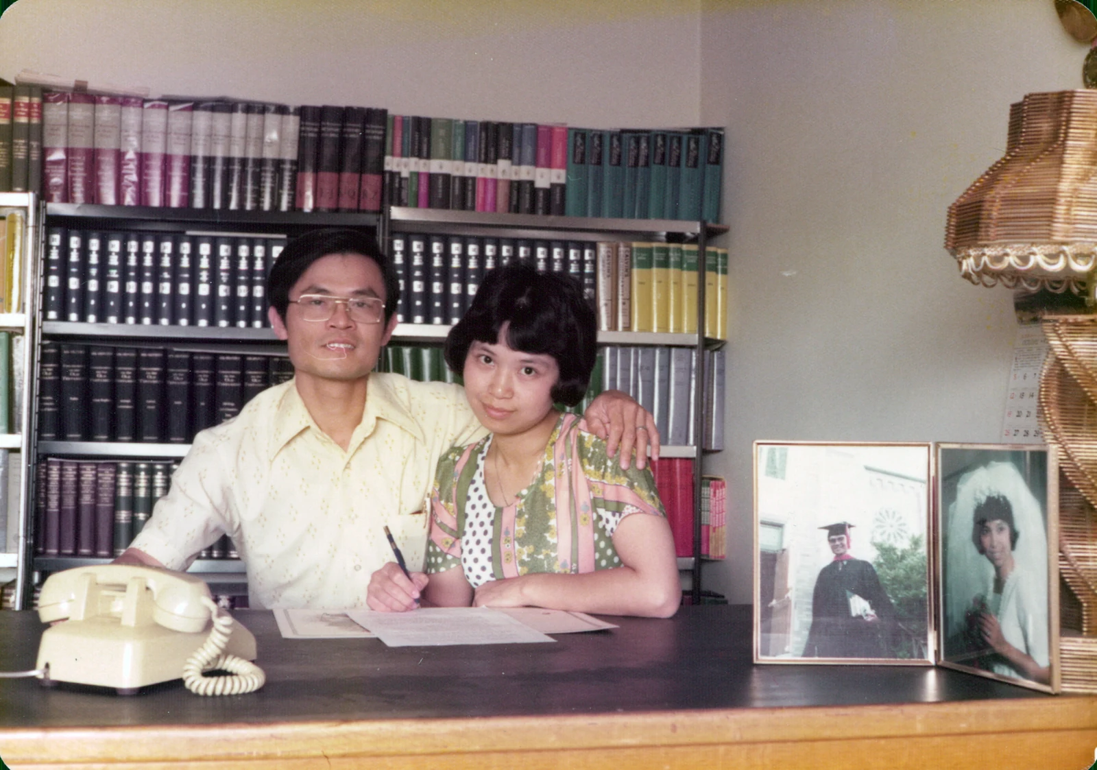 David sits beside Esther with his arm around her shoulders; she holds a pen over papers, with bookshelves behind them.