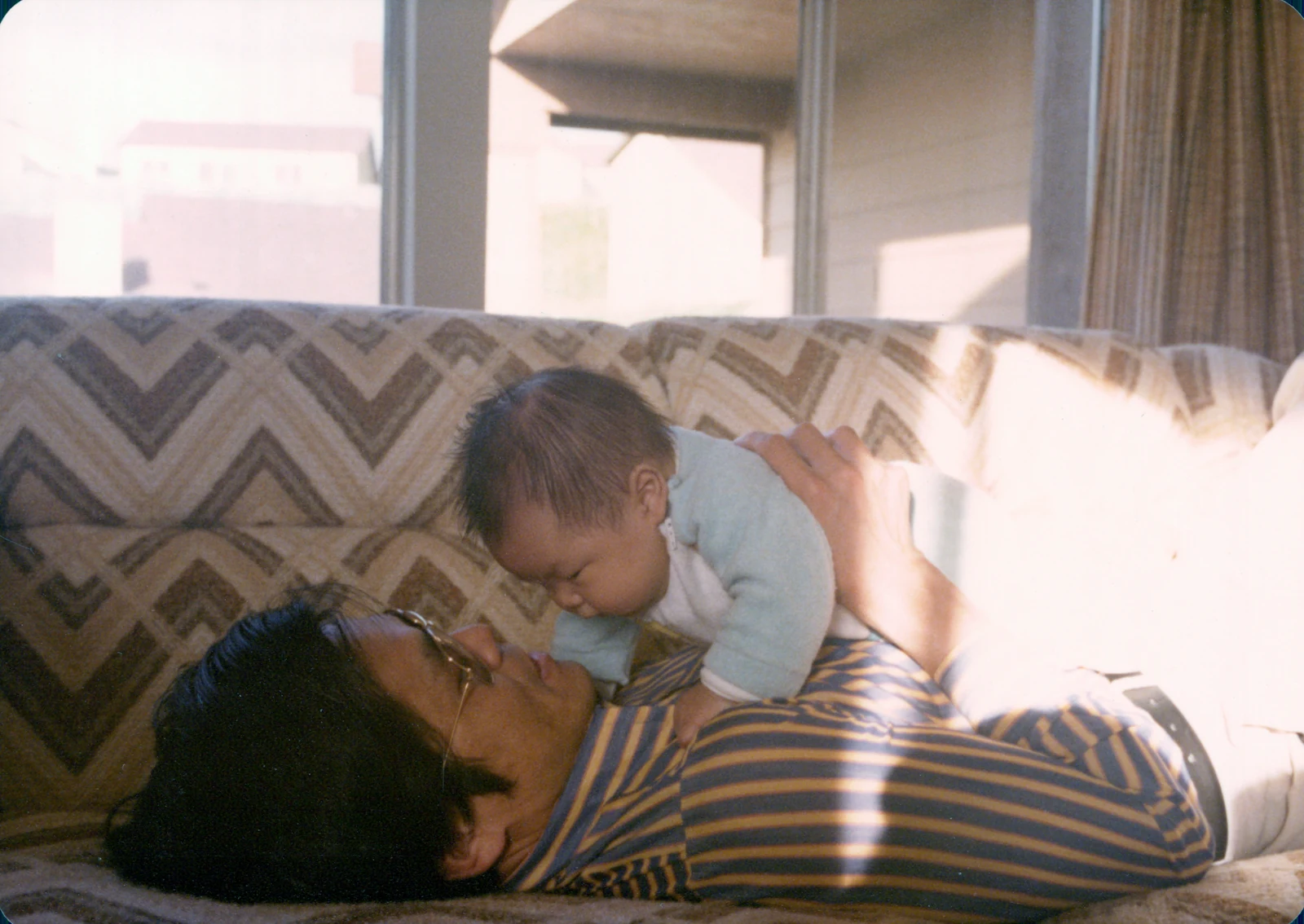 David reclining on a patterned sofa, holding baby Angell up above him.