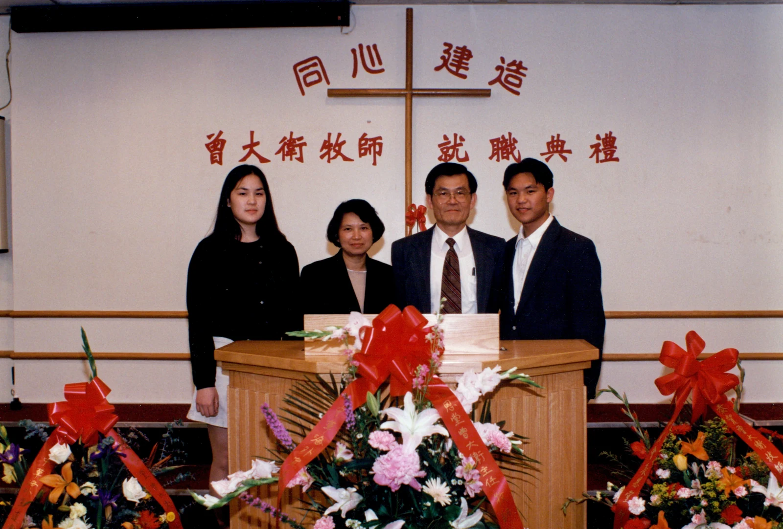 Four people stand behind a flower-covered lectern beneath a cross and Chinese lettering naming Rev. David Tsang’s installation ceremony.