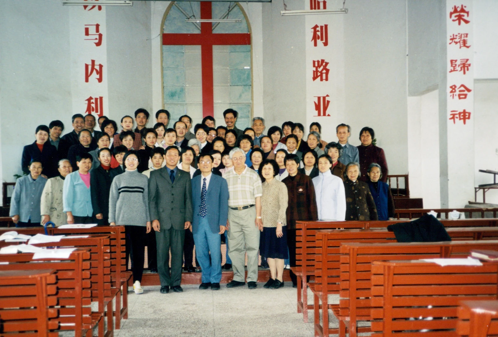 A group poses among wooden church pews beneath a large red cross and Chinese wall lettering.