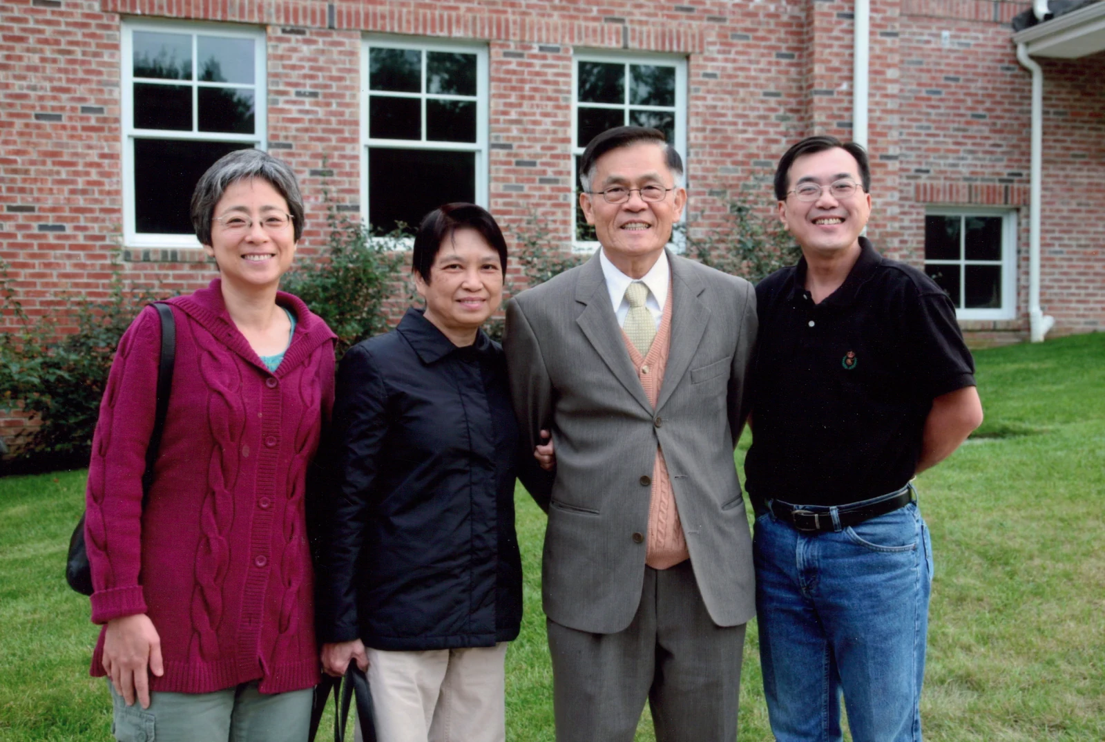 Four people stand together on a lawn outside a brick building with white-framed windows.